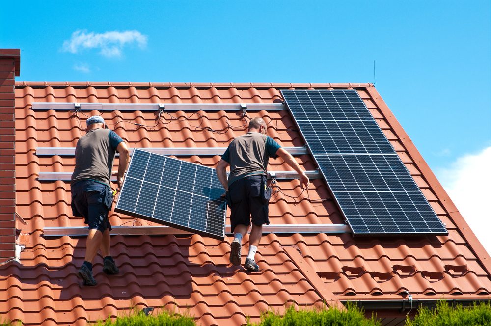 Two installers carrying a solar panel onto rails on a red tile roof, with finished panels beside them
